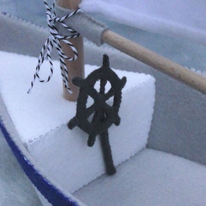 In this picture: a detail of a felt handmade sailing boat,  in white with soft grey and dark blue, with a dark blue steering wheel. The sail is white with bleu lettern WFW.The boat is standing on en soft bleu surface. This boat is made with a PDF handsewing pattern from WoolFeltWonders and is part of a series with felt toy animals ad costumes to portray a profession or hobby. This boat is lovely in combination with some toy animals and the sailor clothing, also a PDF pattern in this series.