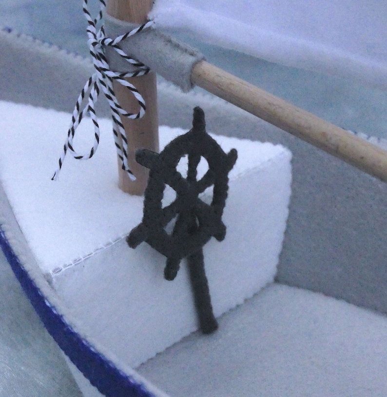 In this picture: a detail of a felt handmade sailing boat,  in white with soft grey and dark blue, with a dark blue steering wheel. The sail is white with bleu lettern WFW.The boat is standing on en soft bleu surface. This boat is made with a PDF handsewing pattern from WoolFeltWonders and is part of a series with felt toy animals ad costumes to portray a profession or hobby. This boat is lovely in combination with some toy animals and the sailor clothing, also a PDF pattern in this series.