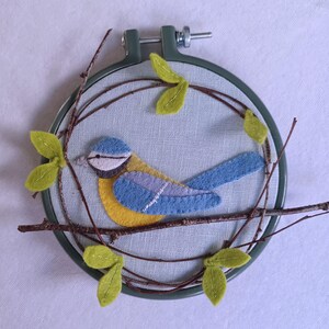 In this picture:  hand made wall decoration with a felt birds on linen in an embroidery hoop, the bird is a blue tit. The hoop is lying on a white surface. These easy to make wall decoration is made with a PDF hand sewing pattern from WoolFeltWonders by Tintangel.