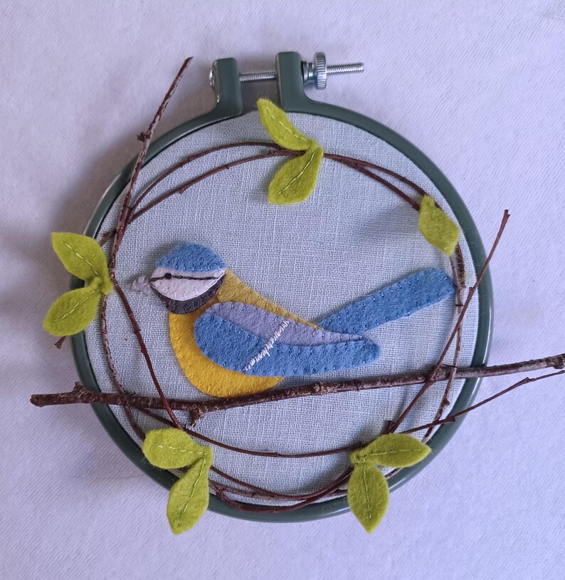 In this picture:  hand made wall decoration with a felt birds on linen in an embroidery hoop, the bird is a blue tit. The hoop is lying on a white surface. These easy to make wall decoration is made with a PDF hand sewing pattern from WoolFeltWonders by Tintangel.