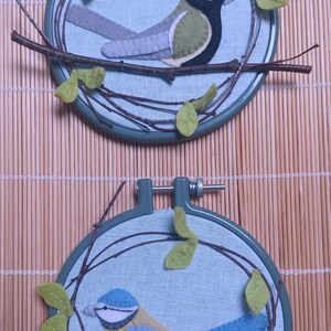 In this picture: two hand made wall decorations with felt birds on linen in an embroidery hoop, one Blue tit and one Great tit. The hoops are lying on a reed mat and some leaves above and besides them. These easy to make wall decorations are made with a PDF hand sewing pattern from WoolFeltWonders by Tintangel.
