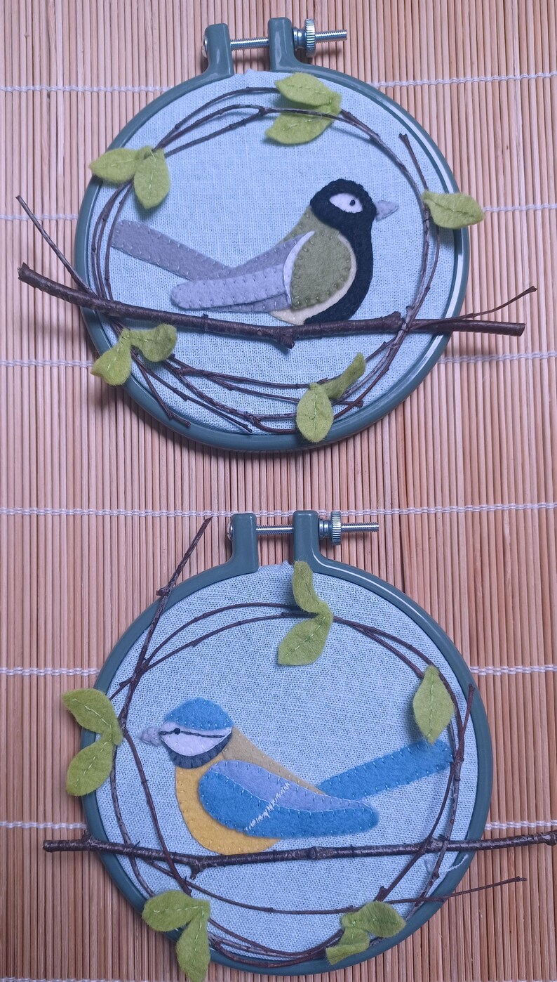 In this picture: two hand made wall decorations with felt birds on linen in an embroidery hoop, one Blue tit and one Great tit. The hoops are lying on a reed mat and some leaves above and besides them. These easy to make wall decorations are made with a PDF hand sewing pattern from WoolFeltWonders by Tintangel.