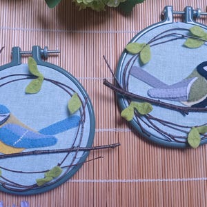 In this picture: two hand made wall decorations with felt birds on linen in an embroidery hoop, one Blue tit and one Great tit. The hoops are lying on a reed mat and some leaves above and besides them. These easy to make wall decorations are made with a PDF hand sewing pattern from WoolFeltWonders by Tintangel.