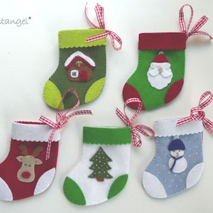 Könnte beinhalten: Fünf Filz-Weihnachtsstrümpfe mit verschiedenen Designs. Die Strümpfe sind grün, rot, blau und weiß mit verschiedenen weihnachtlichen Applikationen, darunter ein Haus, der Weihnachtsmann, ein Rentier, ein Weihnachtsbaum und ein Schneemann.