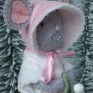 In this picture: a sweet handmade felt mouse with pink dress, white cape and pink and white bonnet, holding a bunch of felt mistletoe with white berries between her hands. She is standing on front of two fake trees on a white surface. This sweet grey mouse is made with a PDF hand sewing pattern from WoolFeltWonders by Tintangel.