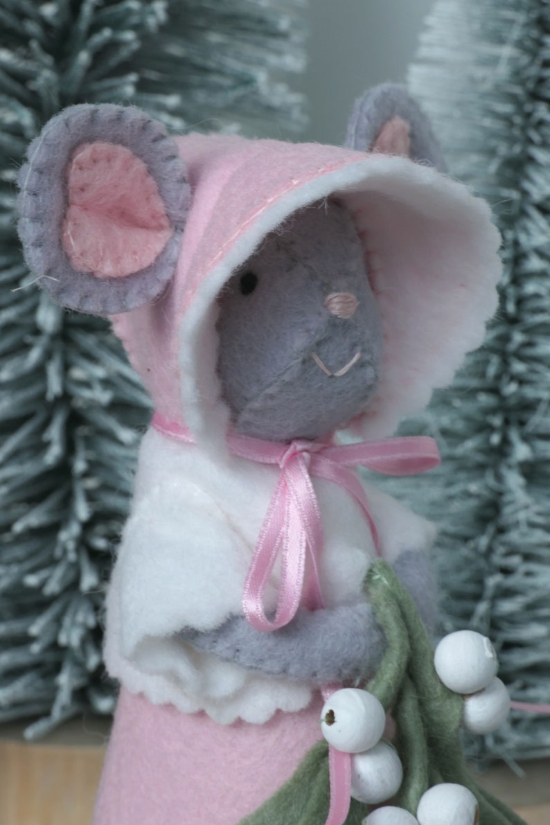 In this picture: a sweet handmade felt mouse with pink dress, white cape and pink and white bonnet, holding a bunch of felt mistletoe with white berries between her hands. She is standing on front of two fake trees on a white surface. This sweet grey mouse is made with a PDF hand sewing pattern from WoolFeltWonders by Tintangel.