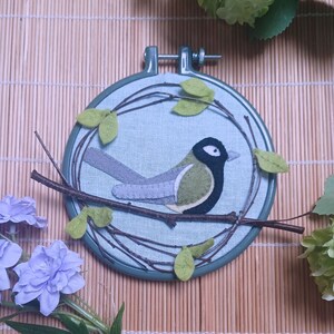 In this picture:  hand made wall decoration with a felt birds on linen in an embroidery hoop, the bird is a Great tit. The hoop is lying on a reed mat and some leaves above and besides it. These easy to make wall decoration is made with a PDF hand sewing pattern from WoolFeltWonders by Tintangel.
