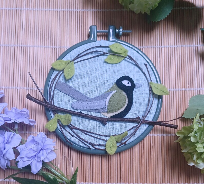 In this picture:  hand made wall decoration with a felt birds on linen in an embroidery hoop, the bird is a Great tit. The hoop is lying on a reed mat and some leaves above and besides it. These easy to make wall decoration is made with a PDF hand sewing pattern from WoolFeltWonders by Tintangel.