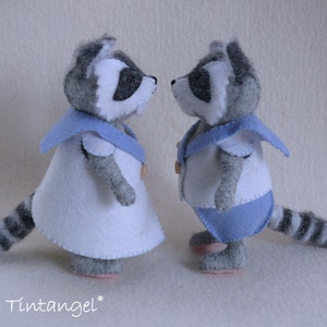 Puede incluir: Dos juguetes de peluche de mapache con atuendos de marinero. El mapache de la izquierda lleva un vestido blanco con cuello azul y una banda azul. El mapache de la derecha lleva una camisa blanca con cuello azul y pantalones azules. Ambos mapaches tienen pelaje gris y blanco y colas rayadas de blanco y negro.