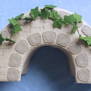 In this picture: a felt handmade  bridge, old style, with ivy vines along the upper side. The bridge is decorated with a stone pattern and is standing on two green waterfronts above blue water. This bridge is part of a Summer sewing project with elegant mice dressed in vintage clothing, Father, mother and two children in a boat.  All made of wool felt with a PDF hand sewing pattern from WoolFeltwonders.