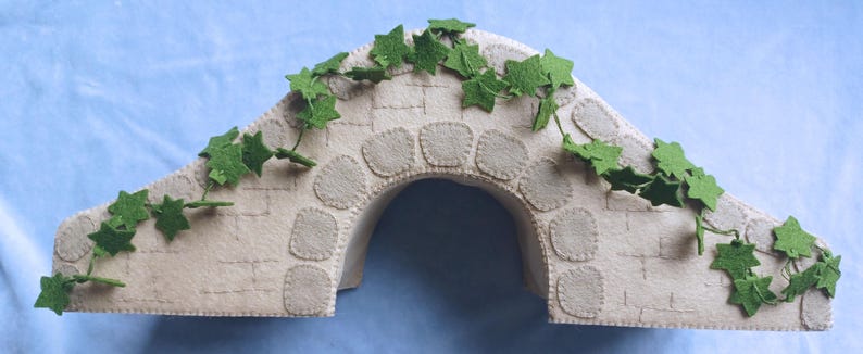 In this picture: a felt handmade  bridge, old style, with ivy vines along the upper side. The bridge is decorated with a stone pattern and is standing on two green waterfronts above blue water. This bridge is part of a Summer sewing project with elegant mice dressed in vintage clothing, Father, mother and two children in a boat.  All made of wool felt with a PDF hand sewing pattern from WoolFeltwonders.