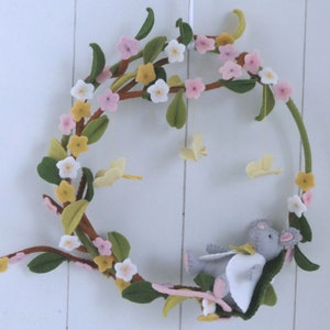 Könnte beinhalten: Ein Filzkranz mit einer grauen Maus, die auf grünen Blättern ruht. Der Kranz ist mit rosa, weißen und gelben Blumen und gelben Schmetterlingsakzenten verziert. Der Kranz wird von einer weißen Schnur vor einem weißen Hintergrund aufgehängt.