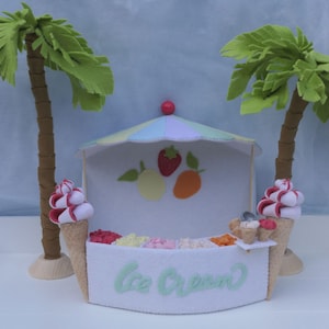 Puede incluir: Un puesto de helados de fieltro con un toldo a rayas pastel, que presenta una variedad de sabores de helado coloridos. Dos palmeras de fieltro flanquean el puesto, y el letrero dice "Ice Cream" en escritura verde. Dos conos de helado con remolinos rojos y blancos a cada lado.