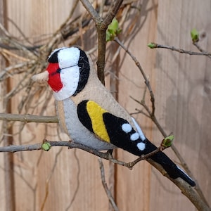 Könnte beinhalten: Ein Filzvogel-Anhänger in Form eines Goldfinken. Der Vogel sitzt auf einem Ast und hat einen roten Kopf, schwarz-weiße Flügel und eine gelbe Brust.