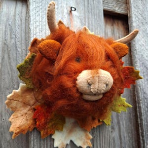 Op de afbeelding: Een naaldgevilte Highland-koeienkop als decoratie met lang, ruig, roestkleurig wol, twee lichtbruine hoorns en een beige vilten snuit. De koe is omringd door een decoratieve schikking van vilten herfstbladeren in tinten oranje, rood en groen.