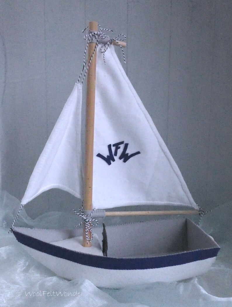 In this picture: a handmade sailing boat, made from felt in white with soft grey and dark blue, with a dark blue steering wheel. The sail is white with bleu lettern WFW.The boat is standing on en soft bleu surface. This boat is made with a PDF handsewing pattern from WoolFeltWonders and is part of a series with felt toy animals ad costumes to portray a profession or hobby. This boat is lovely in combination with some toy animals and the sailor clothing, also a PDF pattern in this series.lauw.