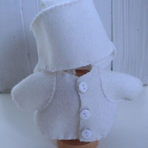 May include: A handmade, white felt doll wearing a chef's hat and jacket with three buttons. The doll is mounted on a small wooden base. The doll's design includes a collar and small, rounded sleeves. The word "Tintangel" is printed at the bottom.