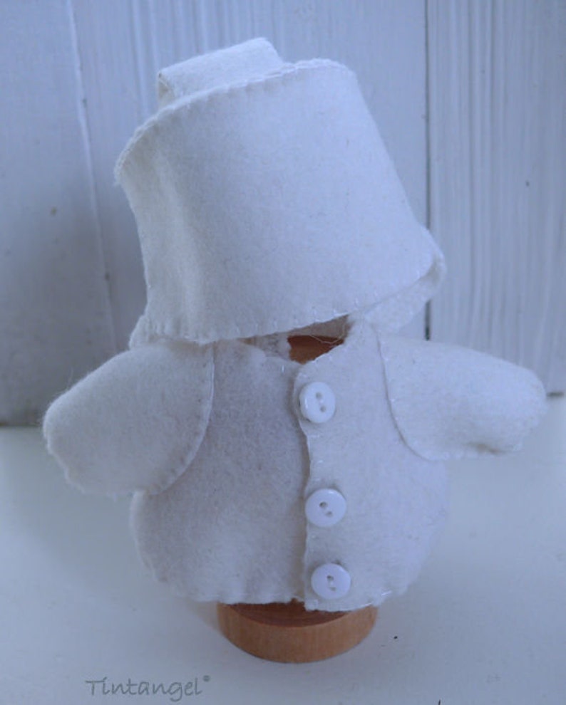 May include: A handmade, white felt doll wearing a chef's hat and jacket with three buttons. The doll is mounted on a small wooden base. The doll's design includes a collar and small, rounded sleeves. The word "Tintangel" is printed at the bottom.