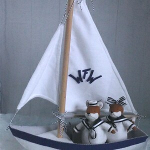 In this picture: a handmade sailing boat, made from felt in white with soft grey and dark blue, with a dark blue steering wheel. The sail is white with bleu lettern WFW. In  the boat two felt toy foxes in sailor costumes. This boat, the foxes and their clothing is made with a PDF handsewing pattern from WoolFeltWonders and is part of a series with felt toy animals ad costumes to portray a profession or hobby.