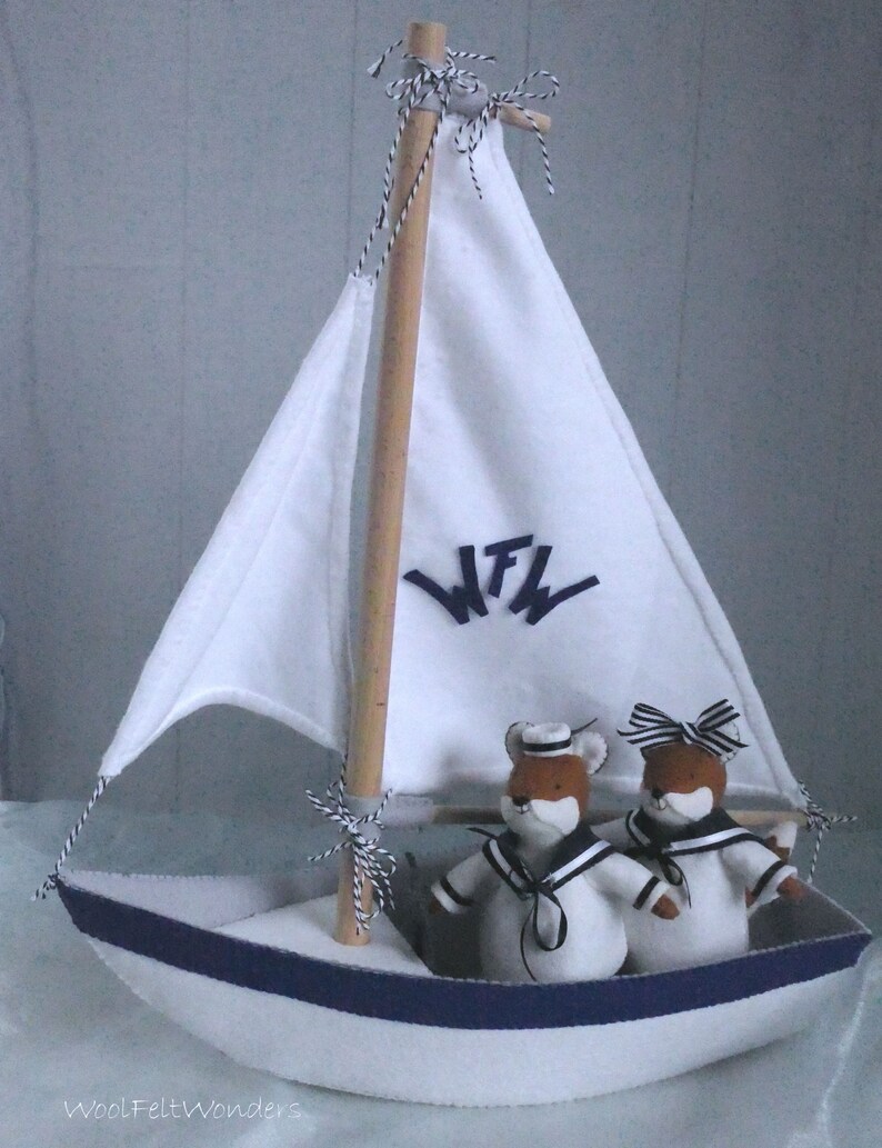 In this picture: a handmade sailing boat, made from felt in white with soft grey and dark blue, with a dark blue steering wheel. The sail is white with bleu lettern WFW. In  the boat two felt toy foxes in sailor costumes. This boat, the foxes and their clothing is made with a PDF handsewing pattern from WoolFeltWonders and is part of a series with felt toy animals ad costumes to portray a profession or hobby.