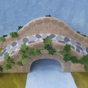 In this picture: a felt handmade  bridge, old style, with ivy vines along the upper side. The bridge is decorated with a stone pattern and is standing on two green waterfronts above blue water. This bridge is part of a Summer sewing project with elegant mice dressed in vintage clothing, Father, mother and two children in a boat.  All made of wool felt with a PDF hand sewing pattern from WoolFeltwonders.