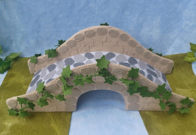 In this picture: a felt handmade  bridge, old style, with ivy vines along the upper side. The bridge is decorated with a stone pattern and is standing on two green waterfronts above blue water. This bridge is part of a Summer sewing project with elegant mice dressed in vintage clothing, Father, mother and two children in a boat.  All made of wool felt with a PDF hand sewing pattern from WoolFeltwonders.