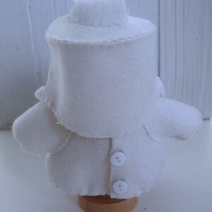 May include: A handmade, white felt doll with a hat, coat, and two buttons. The doll is mounted on a small wooden base. The doll's design is simple and the stitching is visible. The word "Tintangel" is printed on the base.