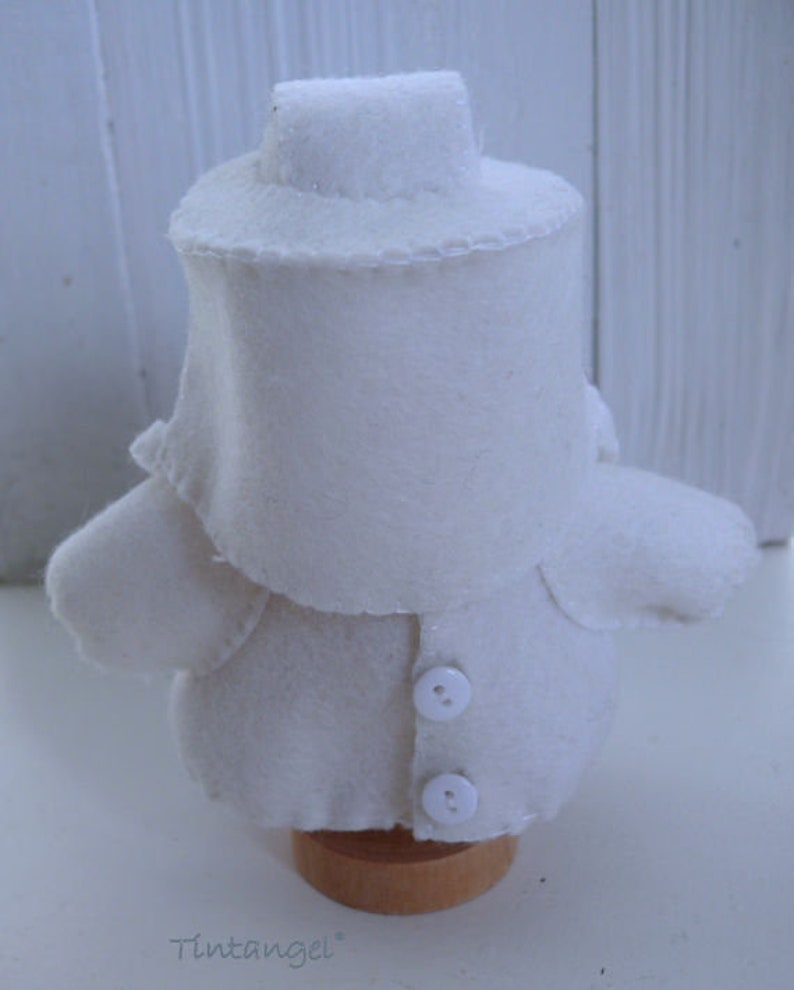 May include: A handmade, white felt doll with a hat, coat, and two buttons. The doll is mounted on a small wooden base. The doll's design is simple and the stitching is visible. The word "Tintangel" is printed on the base.