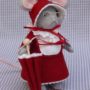 May include: A gray felt mouse wearing a red dress with a white collar and a red felt hat. The mouse is holding a red umbrella with a wooden handle. The mouse is wearing glasses and has a small red bow around its neck.