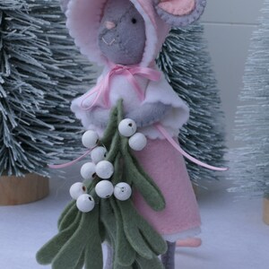 In this picture: a sweet handmade felt mouse with pink dress, white cape and pink and white bonnet, holding a bunch of felt mistletoe with white berries between her hands. She is standing on front of two fake trees on a white surface. This sweet grey mouse is made with a PDF hand sewing pattern from WoolFeltWonders by Tintangel.