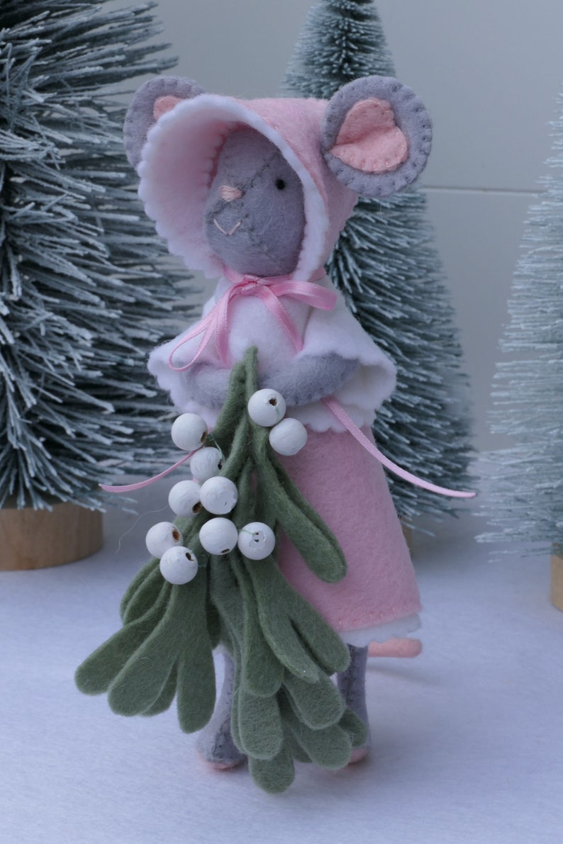 In this picture: a sweet handmade felt mouse with pink dress, white cape and pink and white bonnet, holding a bunch of felt mistletoe with white berries between her hands. She is standing on front of two fake trees on a white surface. This sweet grey mouse is made with a PDF hand sewing pattern from WoolFeltWonders by Tintangel.