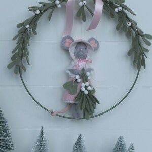 In this picture: a sweet handmade felt mouse with pink dress, white cape and pink and white bonnet, holding a bunch of felt mistletoe with white berries between her hands. She is hanging in a wreath with pink bow and felt mistletoe with white berries, on a white surface. This sweet grey mouse is made with a PDF hand sewing pattern from WoolFeltWonders by Tintangel.