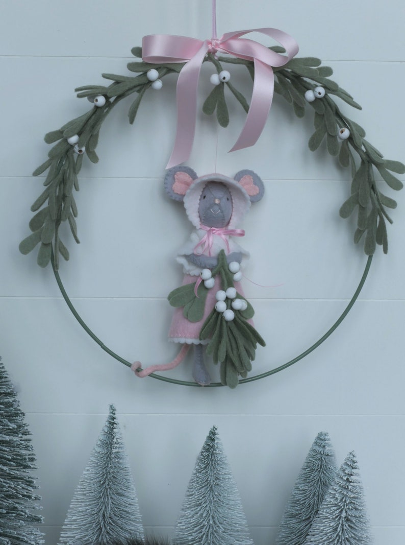 In this picture: a sweet handmade felt mouse with pink dress, white cape and pink and white bonnet, holding a bunch of felt mistletoe with white berries between her hands. She is hanging in a wreath with pink bow and felt mistletoe with white berries, on a white surface. This sweet grey mouse is made with a PDF hand sewing pattern from WoolFeltWonders by Tintangel.