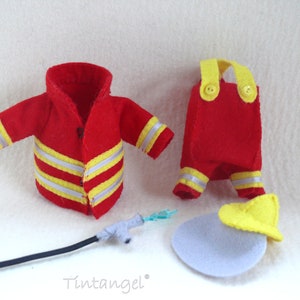 In this picture: a firefigther costumes for felt toy animals, made with a PDF handsewing pattern from WoolFeltWonders, part of a series with toy animal and clothing patterns to mix and match, to portray hobby or profession, for decoration and play. this set has a red jacket and trousers with yellow helmet.  All clothes have reflecting stripes in yellow and silver. A waterhose is lying in front of the clothes.