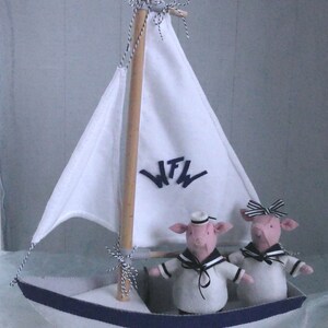 In this picture: a handmade sailing boat, made from felt in white with soft grey and dark blue, with a dark blue steering wheel. The sail is white with bleu lettern WFW. In  the boat two felt toy pigs in sailor costumes. This boat, the pigs and their clothing is made with a PDF handsewing pattern from WoolFeltWonders and is part of a series with felt toy animals ad costumes to portray a profession or hobby.