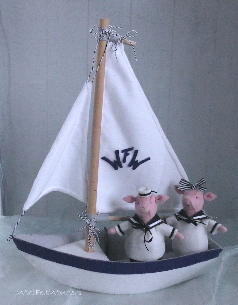 In this picture: a handmade sailing boat, made from felt in white with soft grey and dark blue, with a dark blue steering wheel. The sail is white with bleu lettern WFW. In  the boat two felt toy pigs in sailor costumes. This boat, the pigs and their clothing is made with a PDF handsewing pattern from WoolFeltWonders and is part of a series with felt toy animals ad costumes to portray a profession or hobby.