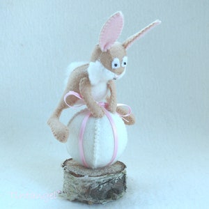 Könnte beinhalten: Ein braunes und weißes Filzkaninchen sitzt auf einem weißen Filzei mit einem rosa Band. Das Ei sitzt auf einem Holzscheit.