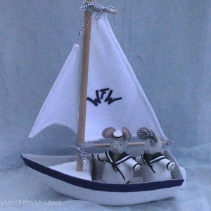 In this picture: a handmade sailing boat, made from felt in white with soft grey and dark blue, with a dark blue steering wheel. The sail is white with bleu lettern WFW. In  the boat two felt toy mice in sailor costumes. This boat, the mice and their clothing is made with a PDF handsewing pattern from WoolFeltWonders and is part of a series with felt toy animals ad costumes to portray a profession or hobby.