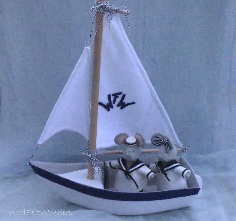 In this picture: a handmade sailing boat, made from felt in white with soft grey and dark blue, with a dark blue steering wheel. The sail is white with bleu lettern WFW. In  the boat two felt toy mice in sailor costumes. This boat, the mice and their clothing is made with a PDF handsewing pattern from WoolFeltWonders and is part of a series with felt toy animals ad costumes to portray a profession or hobby.