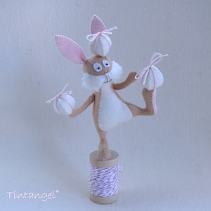 May include: A brown and white felt bunny rabbit with pink ears and a white felt tail. The bunny is balancing on one leg and holding three white felt balls with pink ribbons. The bunny is standing on a spool of red and white twine.