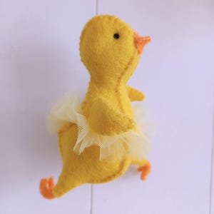 Op de afbeelding: Een handgemaakt geel vilten eendje met een crèmekleurige tule tutu. Het eendje heeft een oranje snavel en voetjes en een zwart oog. Het speelgoed is ontworpen om op een ballerina te lijken.