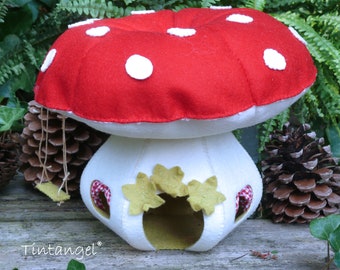 May include: A red and white felt mushroom with white polka dots. The mushroom has a white felt base with a small opening and green felt leaves. The mushroom is sitting on a wooden surface with pine cones.