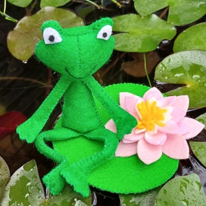 Puede incluir: Una rana de fieltro verde sentada en un nenúfar de fieltro verde con una flor de fieltro rosa. La rana tiene ojos blancos y una amplia sonrisa.