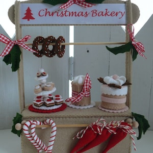 May include: A felt Christmas bakery stand with a sign that reads "Christmas Bakery". The stand is decorated with red and white gingham ribbon, green felt leaves, and a small felt Christmas tree. The stand is filled with felt Christmas treats, including gingerbread cookies, candy canes, and cakes.