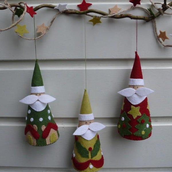 May include: Three cone-shaped felt ornaments hanging from a branch decorated with small felt stars. The left ornament is green with white dots and red house shapes. The center is yellow with green leaves. The right is red with green trees and yellow stars.