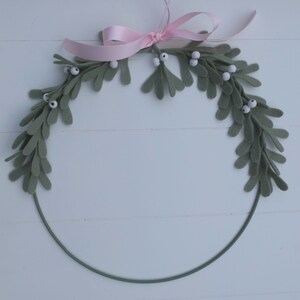 In this picture: a green metal ring on the upperside deocrated with green felt mistletoe with white berries and a pink bow. This ring is part of a design with a mouse, dressed in a pink dress with white cape, white and pink bonnet, holding more Mistletoe between her hands. Made with a PDF hand sewing pattern from WoolFeltwonders by Tintangel.