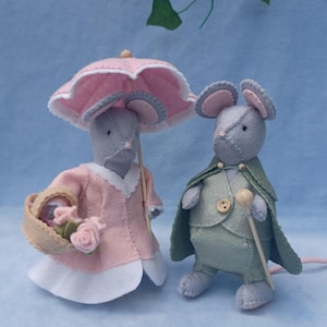 Peut inclure: Deux souris grises en feutre vêtues de costumes d'époque. L'une porte une robe rose, un panier de fleurs et un parasol rose. L'autre porte une cape verte et tient une canne en bois.