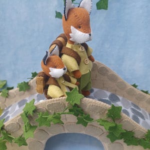 In this picture: a felt handmade  bridge, old style, with ivy vines along the upper side. The bridge is decorated with a stone pattern and is standing on two green waterfronts above blue water. On the bridge a pair of foxes, dressed in green an wearing backpack to go for a hike.  This bridge is part of a Summer sewing project with elegant mice dressed in vintage clothing, Father, mother and two children in a boat.  All made of wool felt with a PDF hand sewing patterns from WoolFeltwonders.