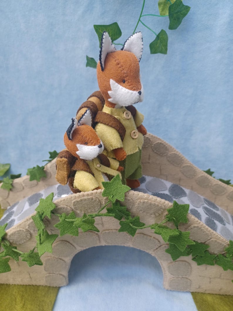 In this picture: a felt handmade  bridge, old style, with ivy vines along the upper side. The bridge is decorated with a stone pattern and is standing on two green waterfronts above blue water. On the bridge a pair of foxes, dressed in green an wearing backpack to go for a hike.  This bridge is part of a Summer sewing project with elegant mice dressed in vintage clothing, Father, mother and two children in a boat.  All made of wool felt with a PDF hand sewing patterns from WoolFeltwonders.