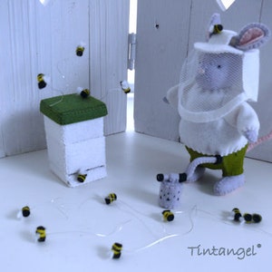 In this picture: a handmade felt mouse dressed as a beekeeper, with white jacket, bee hood, beehive, smoke pipe and many bees. All this is is made with PDF hand sewing pattern from WoolFeltWonders by Tintangel and is part of a series with felt toy animals and all kinds of clothing sets to mix and match to portray hobby or profession. Easy to make animals and clothing sets for decoration and play.
